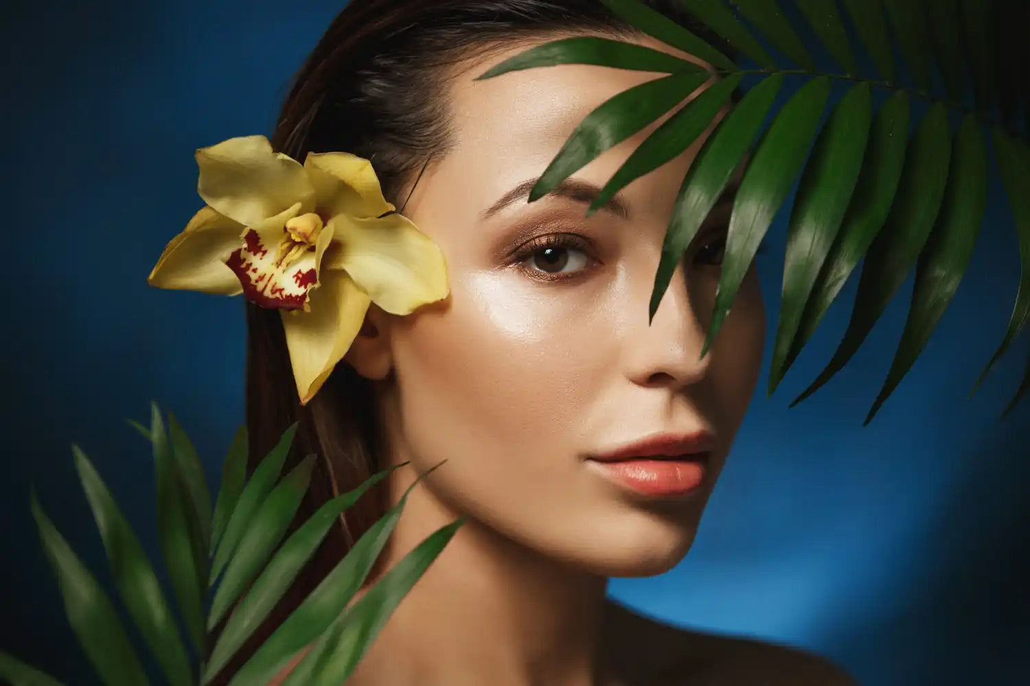 Une femme avec une orchidée jaune dans les cheveux et une peau lumineuse qui dépasse à travers des feuilles tropicales.